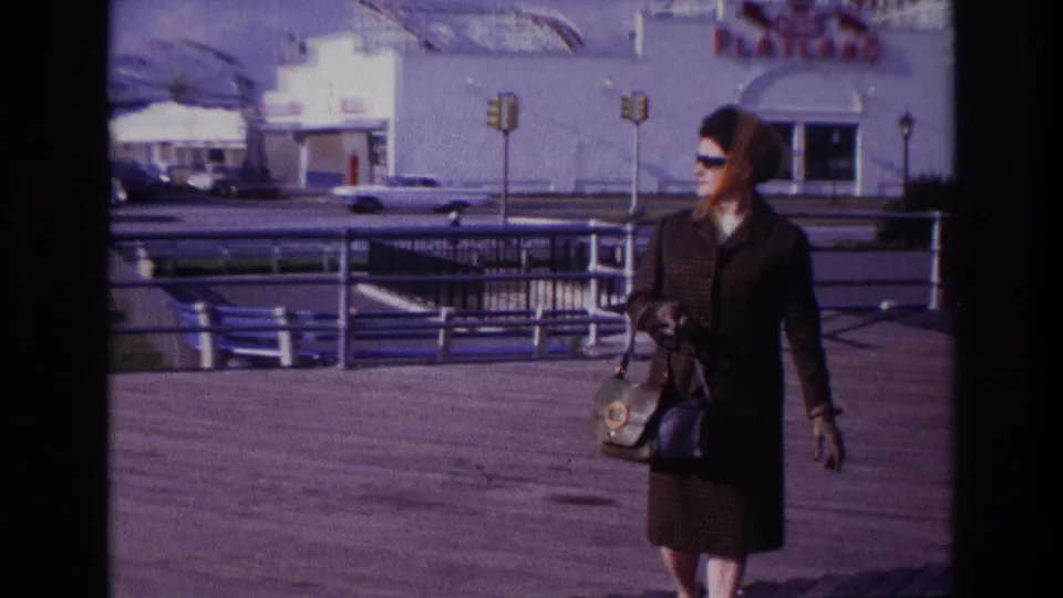 A closer 1967 Rockaway Beach view shows the woman in a headscarf and sunglasses walking past boardwalk railings with traffic and Playland behind.