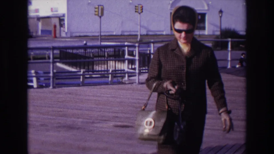Near the end of the clip, the woman fills more of the frame as she carries bags across the Rockaway Beach boardwalk in front of Playland.