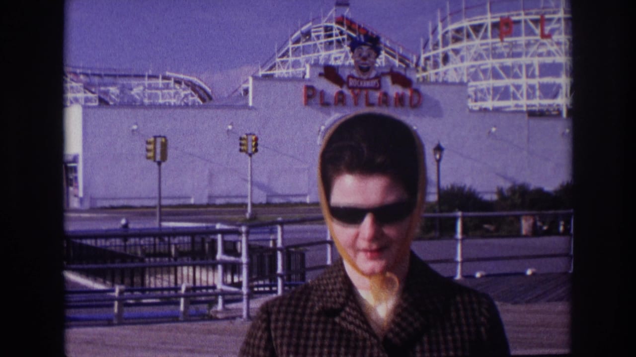 1967 Rockaway Beach home-movie frame showing a woman in sunglasses and yellow headscarf near the Playland sign and roller coaster.