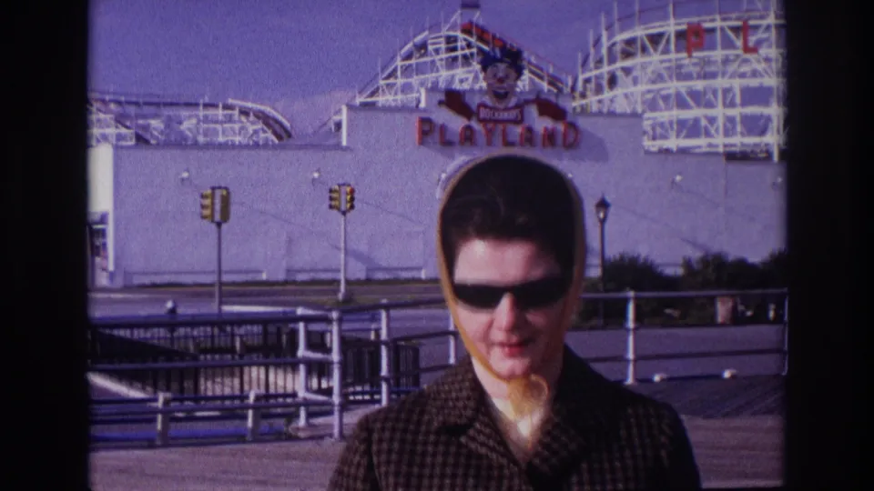 1967 Rockaway Beach home-movie frame showing a woman in sunglasses and yellow headscarf near the Playland sign and roller coaster.
