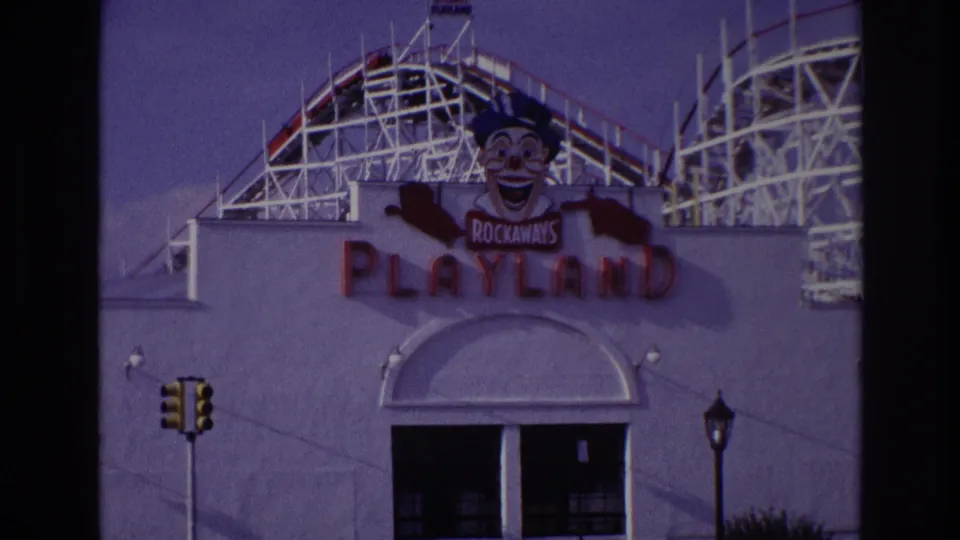 1967 Rockaway Beach frame showing Rockaways Playland signage, clown figure, traffic light, arched entrance, and roller coaster behind the facade.