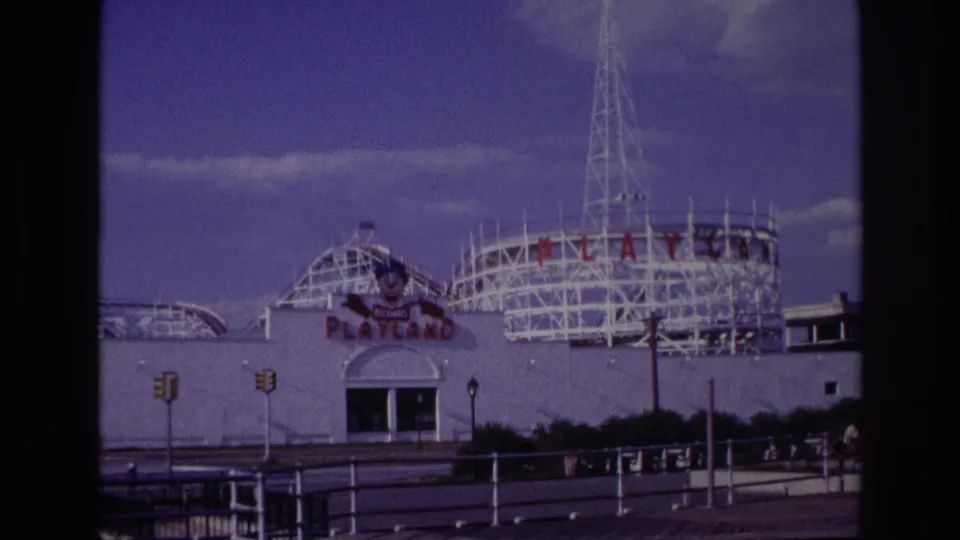 Wider 1967 view of Rockaway Beach Playland showing the entrance facade, white roller-coaster tracks, tall lattice tower, street signals, and foreground railings.