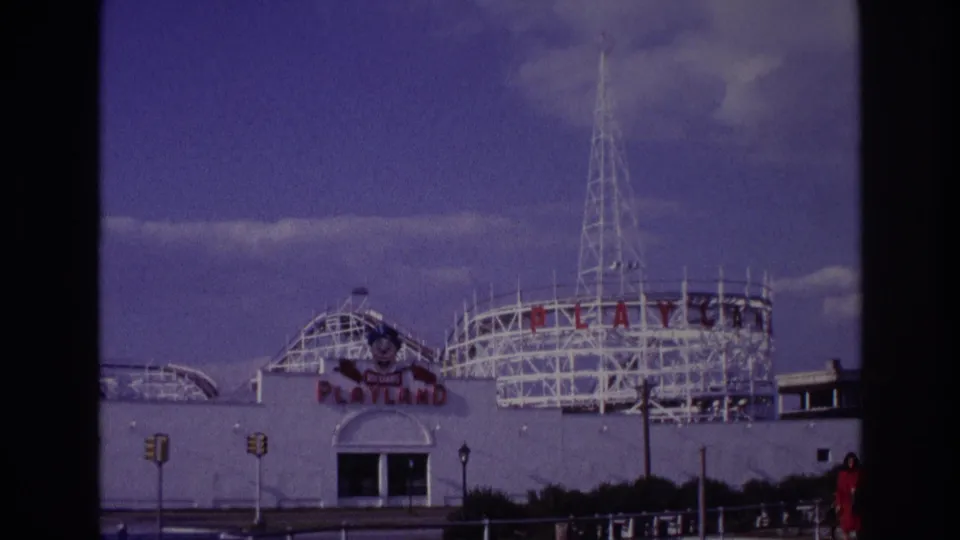 1967 Rockaway Beach Playland facade framed lower in the shot, with white ride structures, a tall tower, traffic lights, and street or boardwalk railings in front.