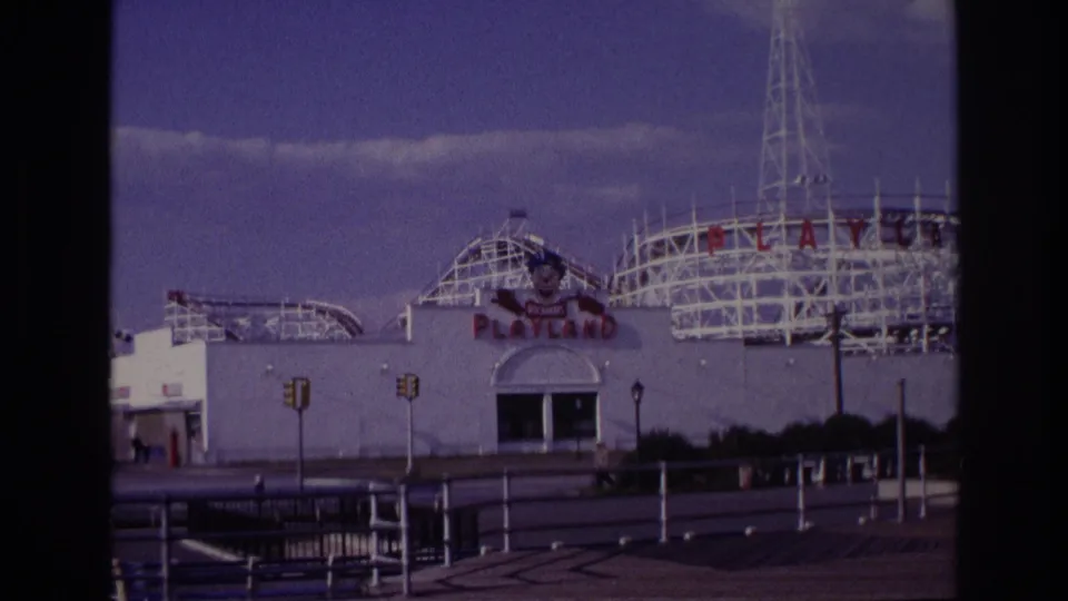 1967 Rockaway Beach Playland entrance and roller-coaster structure seen across pavement and railings, with a small distant pedestrian visible near the lower right edge.