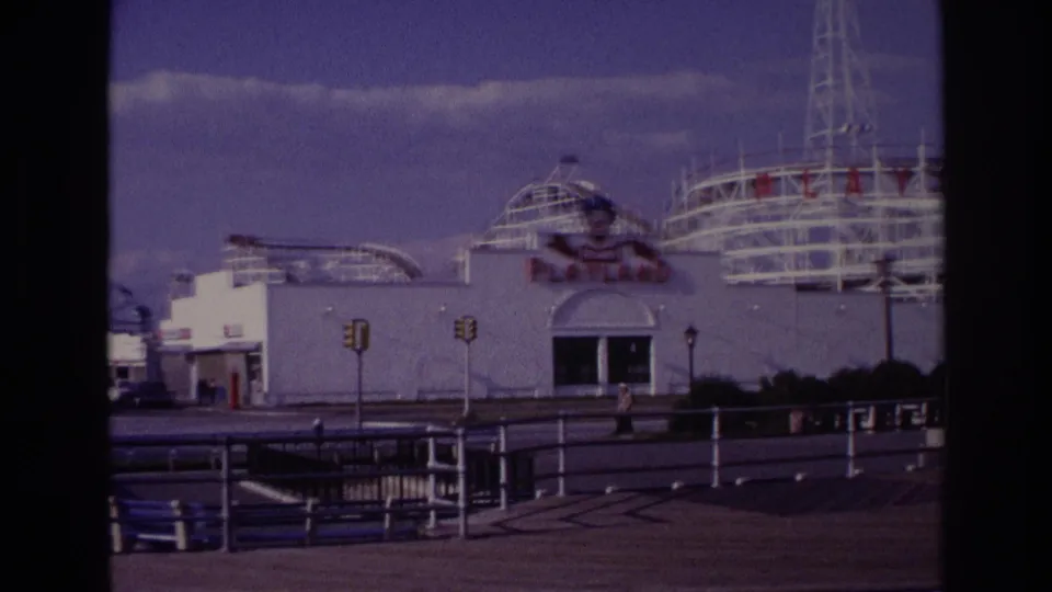 1967 Rockaway Beach Playland exterior with white buildings, rollercoaster structures, traffic lights, railings, benches, and a quiet boardwalk foreground.