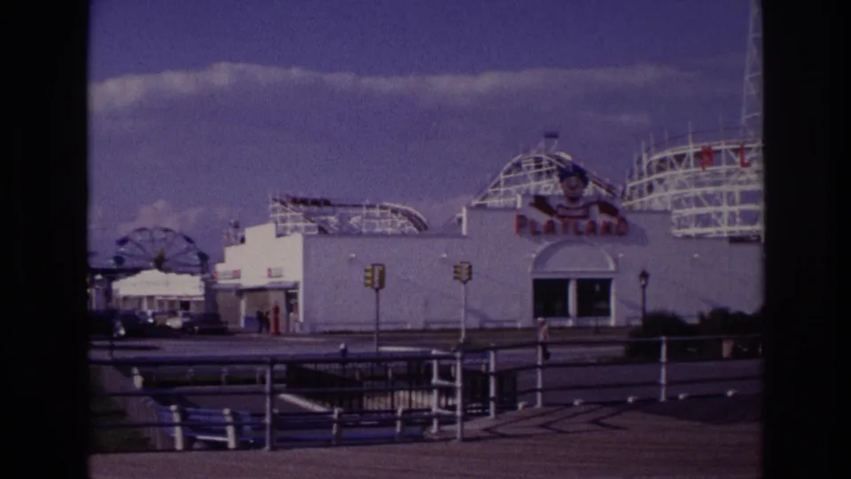 Rockaway Beach Playland exterior in 1967 with the park facade, rollercoaster framework, a distant Ferris wheel, traffic lights, railings, and benches.