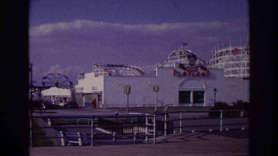 1967 home-movie view of Rockaway Beach Playland, showing the entrance area, rollercoaster tracks, Ferris wheel, boardwalk railings, and pedestrians.