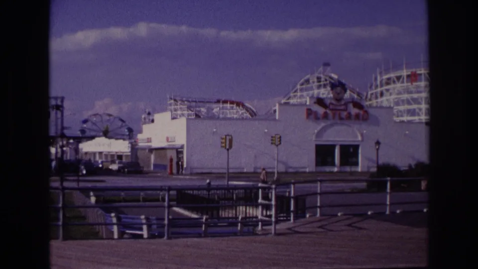 Rockaway Beach Playland exterior at the end of the clip, with rollercoaster tracks, white facade, railings, traffic lights, benches, and a pedestrian nearby.