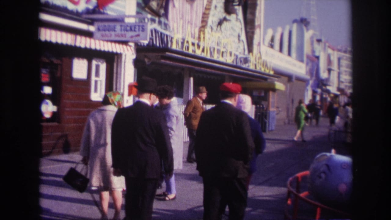 Adults walk along an amusement boardwalk past ticket signage and ride structures.