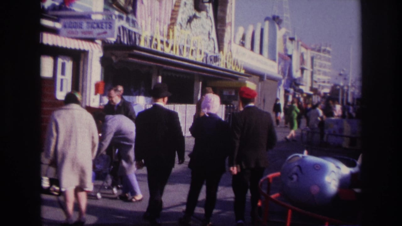 Adults in period clothing walk along the Rockaway Beach amusement boardwalk beside a blue whale-like children’s ride.