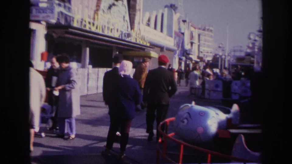 Rockaway Beach boardwalk scene with suited adults, a red-capped man, amusement railings, and a blue children’s ride in the foreground.