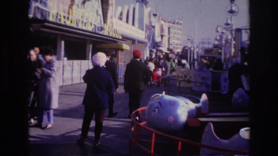 1967 Rockaway Beach amusement boardwalk with adults walking away from camera, ticket railings, lampposts, and a blue ride vehicle.