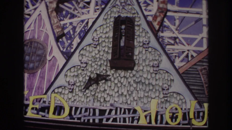 1967 Rockaway Beach frame of a themed facade with a dark figure in the window, skull details, yellow signage, and ride framework.