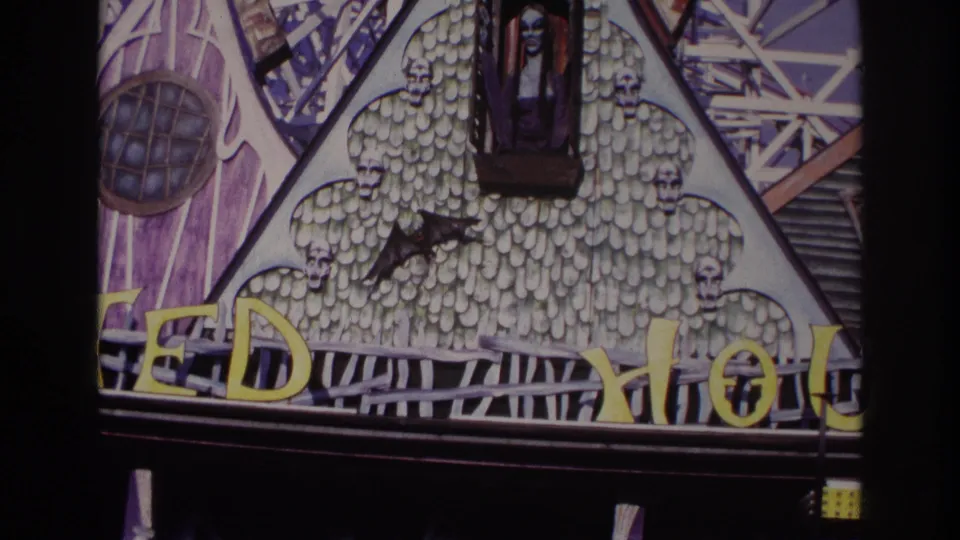 1967 Rockaway Beach home-movie frame of a gothic amusement facade with a window figure, skull trim, bat shape, and yellow letters.