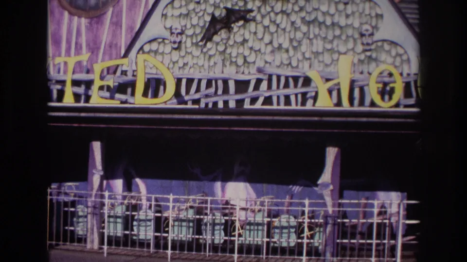 Rockaway Beach 1967 home-movie frame showing the lower TEDDY HOUSE sign area, dark awning, metal railing, and faint silhouettes behind it.