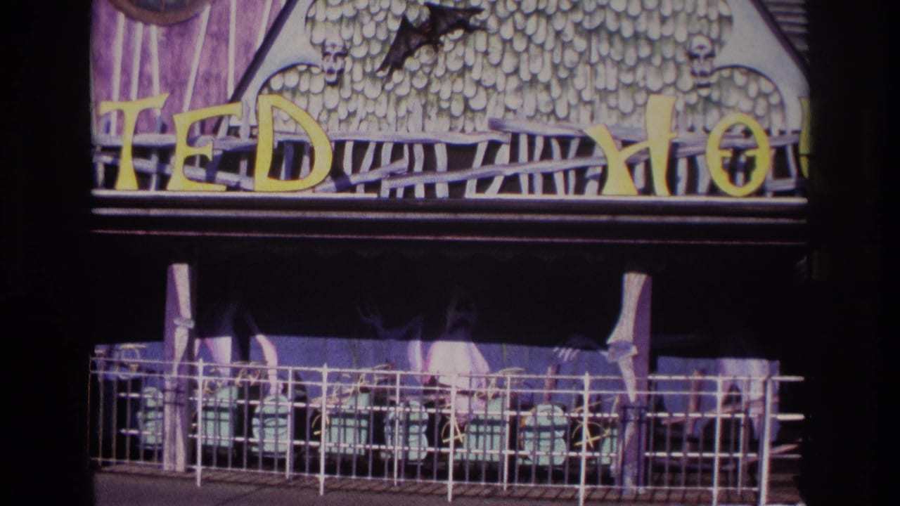 1967 Rockaway Beach 8mm frame showing a haunted-house style facade with skull motifs, a black bat cutout, yellow sign letters, and a railed entrance.