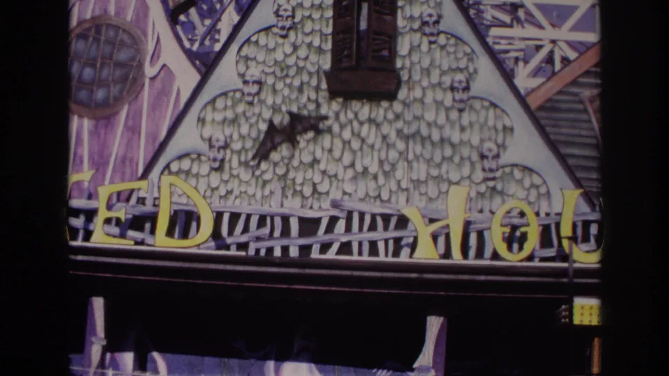 Final 1967 Rockaway Beach frame of the haunted attraction front, showing the bat cutout, skull patterns, partial yellow sign, angled trim, and dark railed entry.