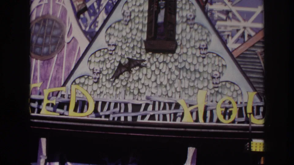 1967 Rockaway Beach home-movie frame showing a colorful haunted house attraction facade with skull motifs, a bat figure, a small ghostly window figure, and partial yellow signage.