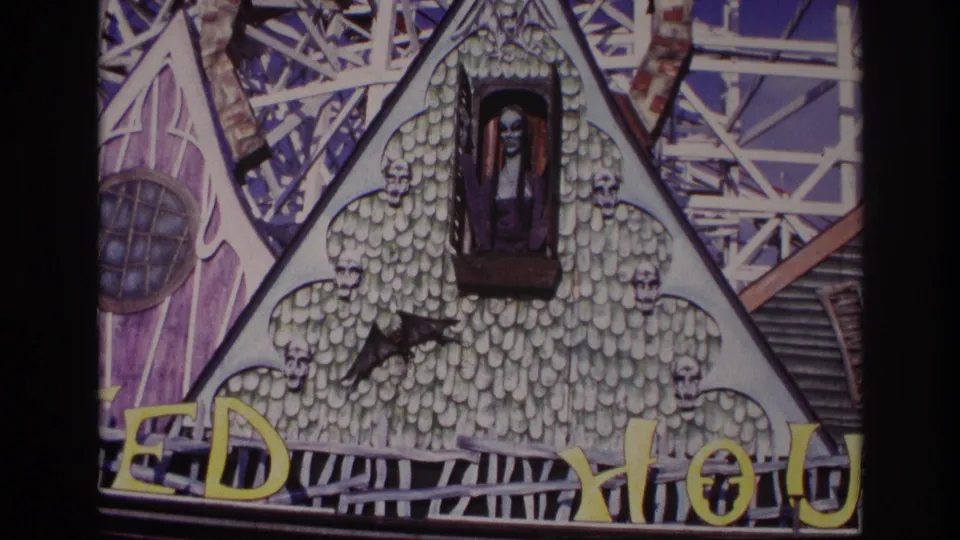1967 Rockaway Beach home-movie frame showing the haunted house facade from a slightly lower framing, including skull motifs, a bat figure, purple trim, and cropped yellow attraction lettering.