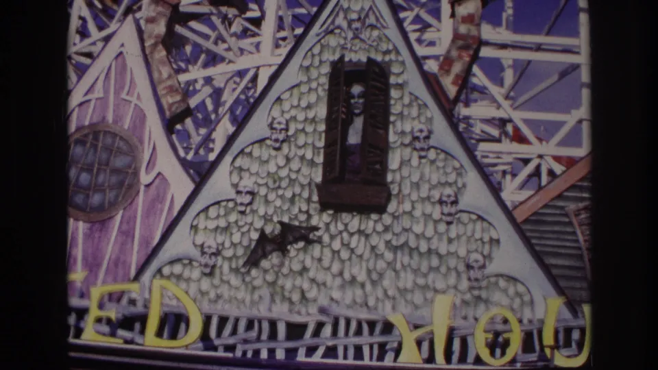 1967 Rockaway Beach frame showing the haunted attraction facade with a skull-covered painted front, bat figure, small window figure, exposed framework, and cropped yellow signage.