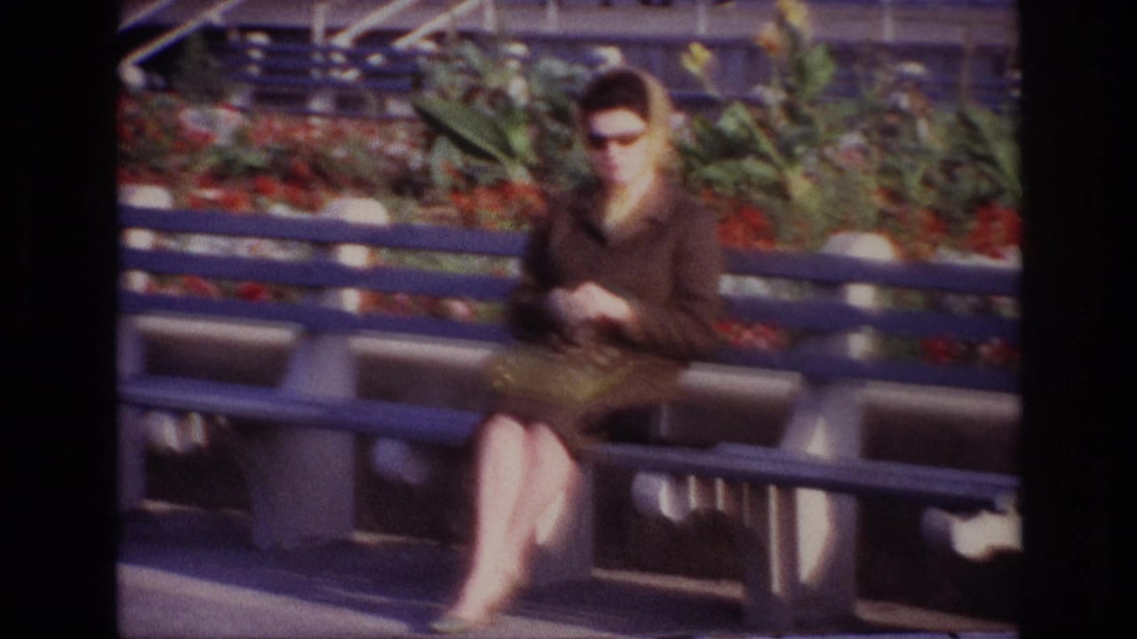 At Rockaway Beach in 1967, a woman in sunglasses and a brown coat sits on a blue bench with red flowers and foliage behind her.