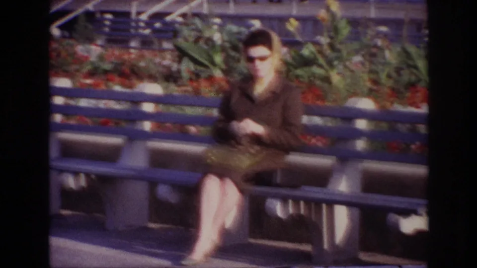 At Rockaway Beach in 1967, a woman in sunglasses and a brown coat sits on a blue bench with red flowers and foliage behind her.