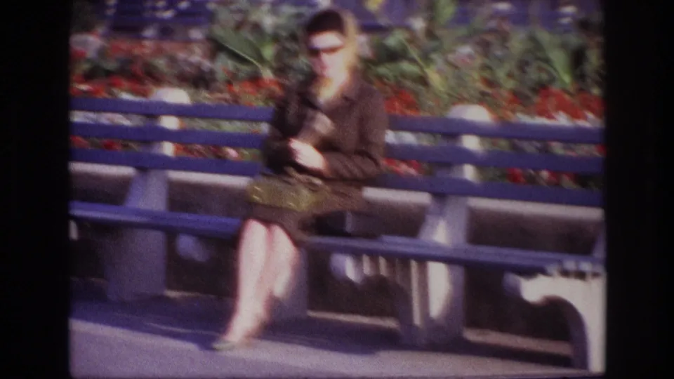 The 1967 Rockaway Beach home-movie frame shows a woman seated cross-legged on a blue bench, handling gloves near a small handbag.