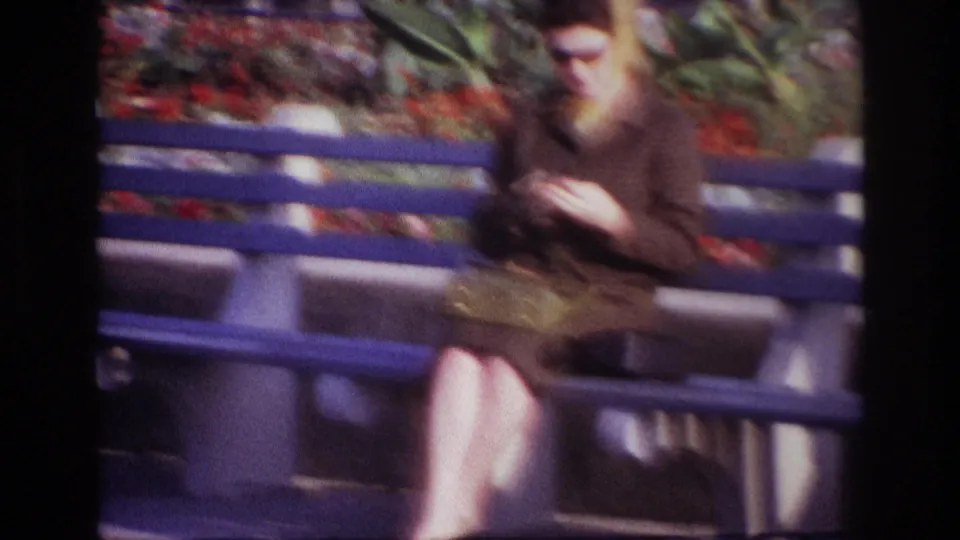 In the final 1967 Rockaway Beach frame, the woman leans over her hands on the bench, with bright red flowers and foliage in the background.