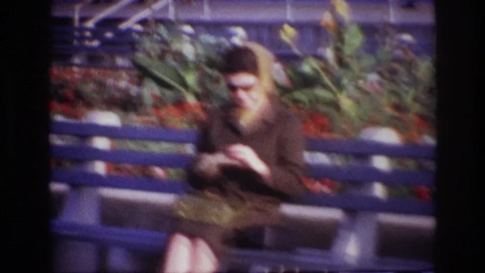 At Rockaway Beach in 1967, a woman in sunglasses sits on a blue bench beside flowers and greenery.