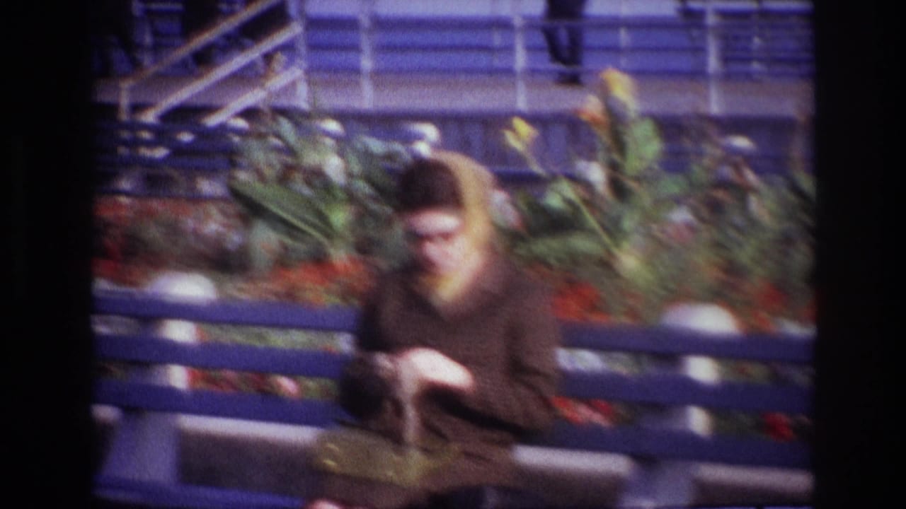 A woman in a brown coat and headscarf sits on a blue bench at Rockaway Beach, looking down at her hands.