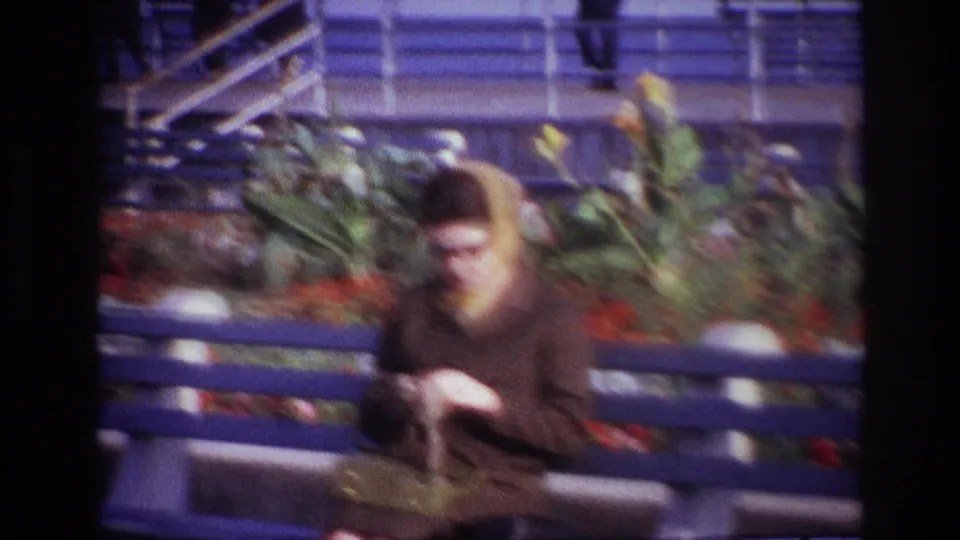 A woman in a brown coat and headscarf sits on a blue bench at Rockaway Beach, looking down at her hands.
