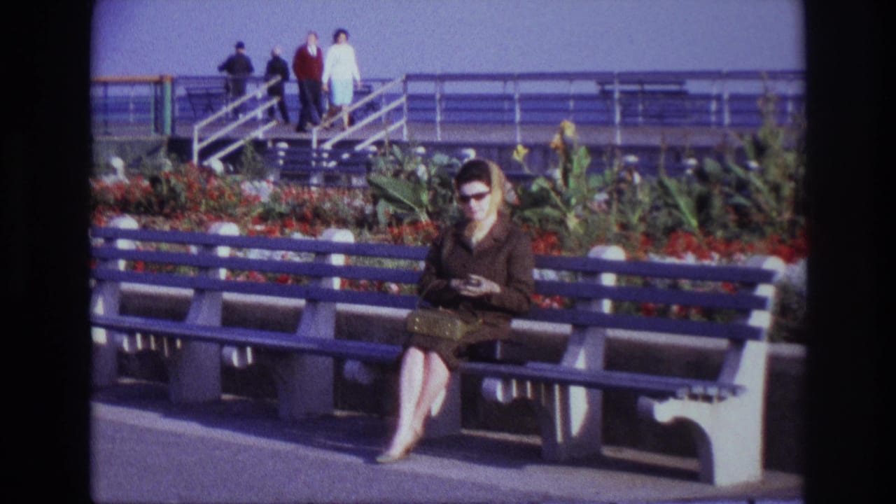 At Rockaway Beach in 1967, a woman in sunglasses and a dark coat sits on a blue bench with flowers, railings and distant walkers behind her.