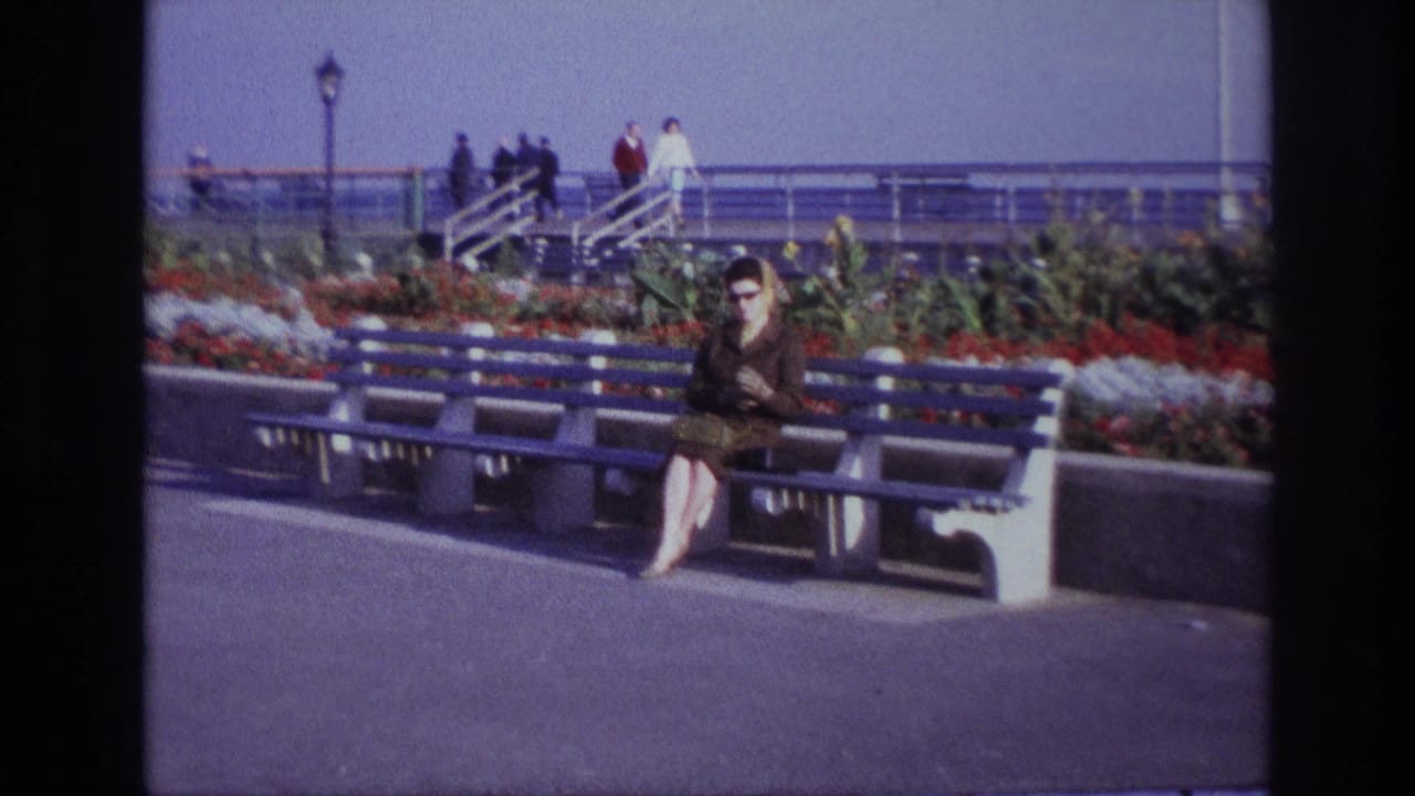 The camera has pulled back slightly on the Rockaway Beach promenade, showing the seated woman, blue benches, flowers, railings and people in the distance.