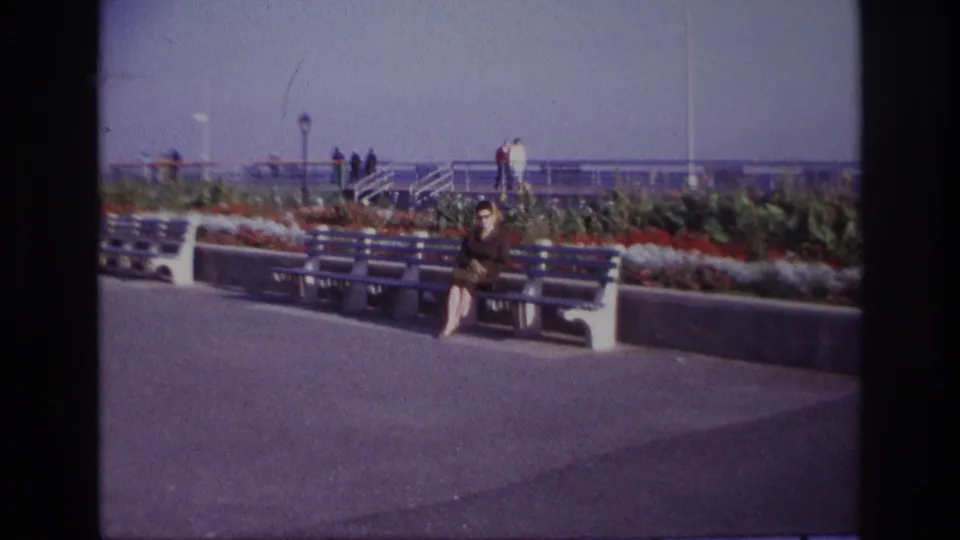 By the end of the pullback, the Rockaway Beach promenade is shown broadly, with the seated woman, benches, flower beds, railings and distant visitors.