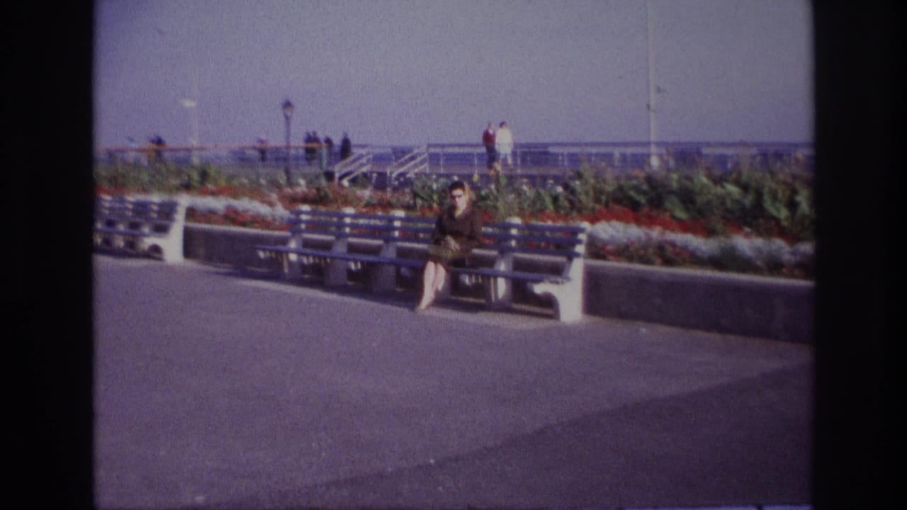 At Rockaway Beach in 1967, a woman sits on a blue and white bench near flowers, railings and distant visitors.