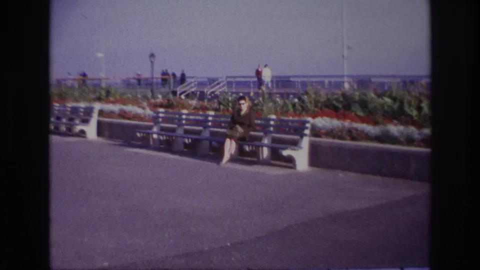 At Rockaway Beach in 1967, a woman sits on a blue and white bench near flowers, railings and distant visitors.