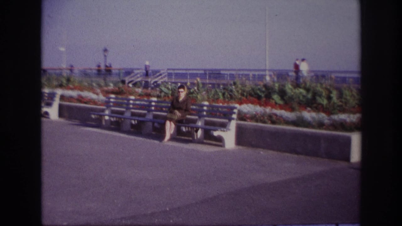 Woman seated on a bench as the camera begins to zoom toward her from the promenade.