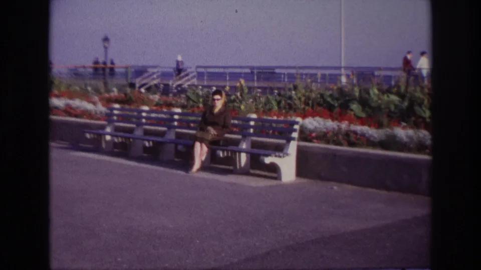 At Rockaway Beach in 1967, the zoom brings the seated woman nearer while the planted bed and waterfront railings remain visible.