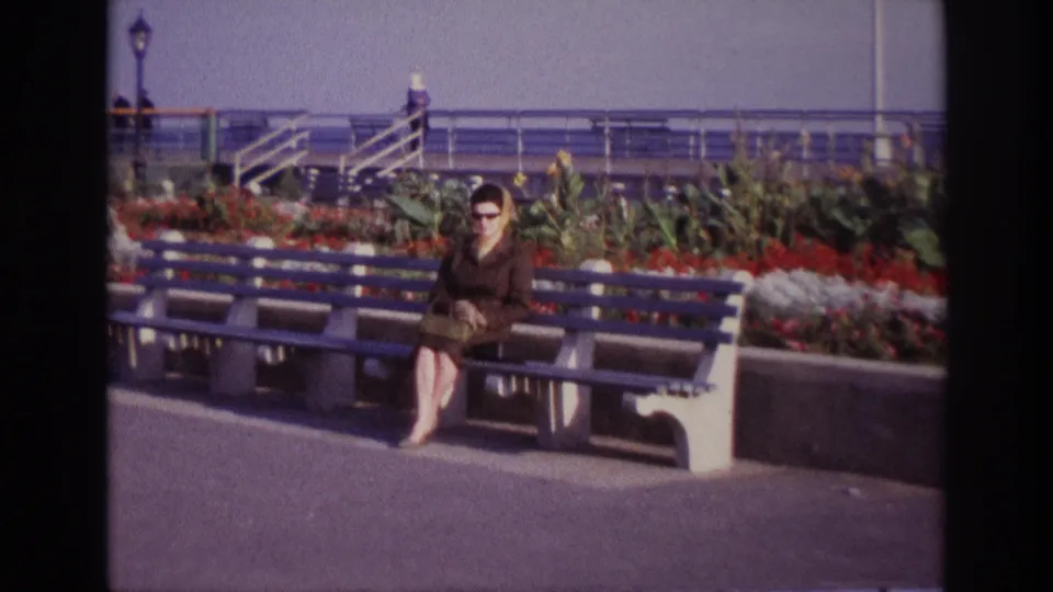 The zoom ends on the woman seated at Rockaway Beach in 1967, with blue bench slats, planted flowers and waterfront railings behind.