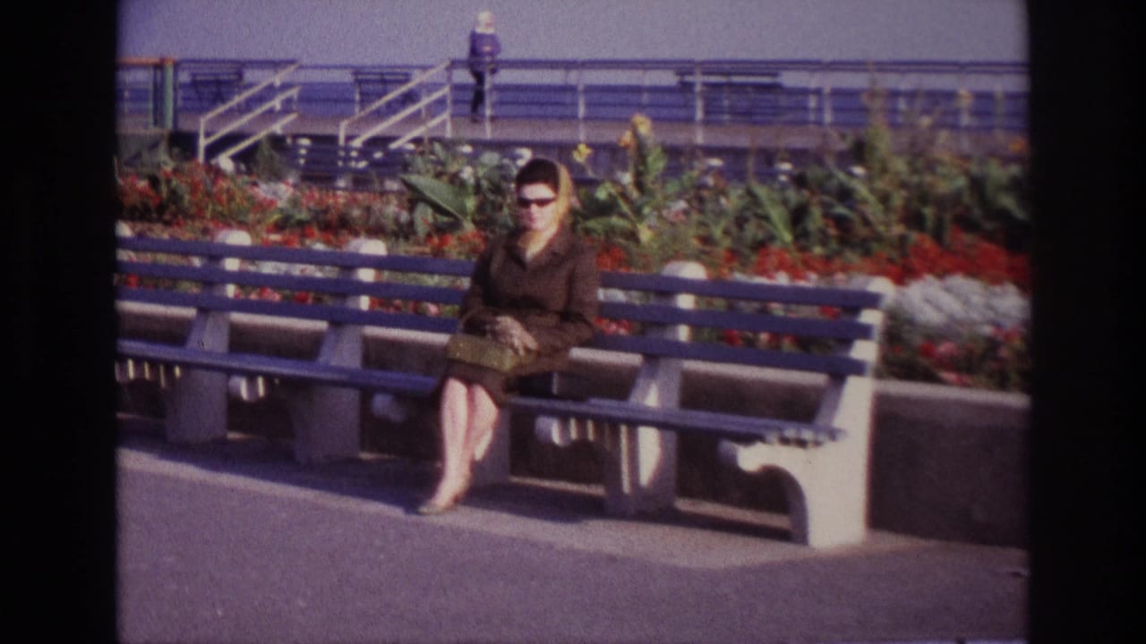 At Rockaway Beach in 1967, a woman in a brown coat and sunglasses sits on a blue bench near a flower bed and railings.