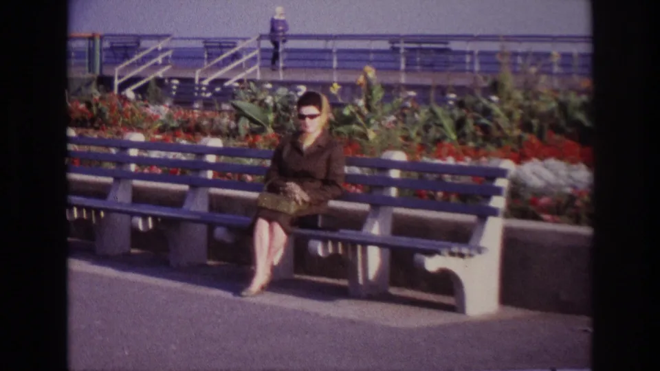 At Rockaway Beach in 1967, a woman in a brown coat and sunglasses sits on a blue bench near a flower bed and railings.