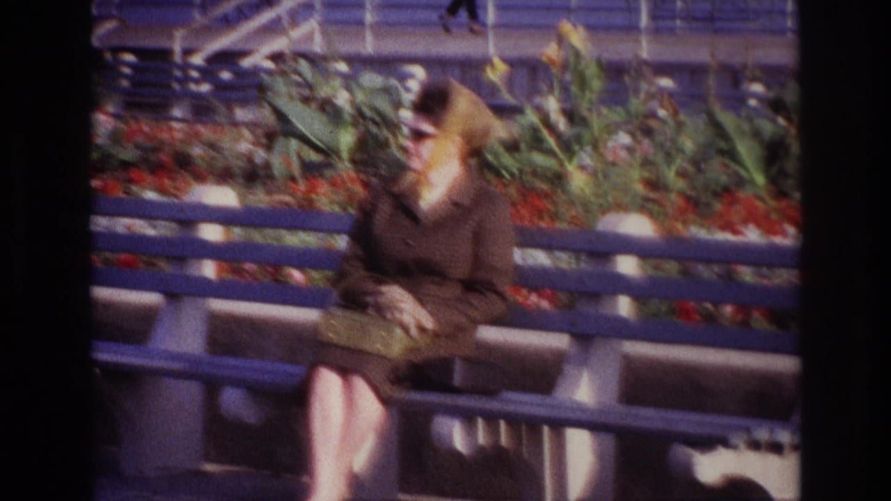 Closer view of a woman in a headscarf and sunglasses seated on a bench with flowers behind her.