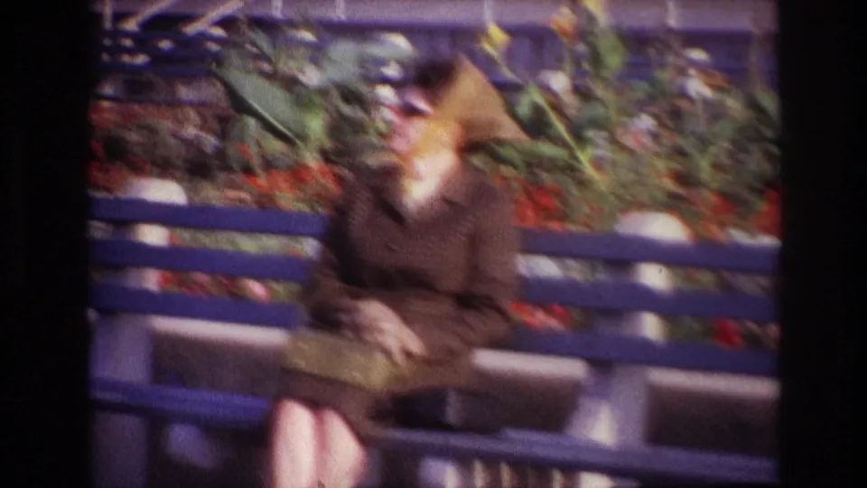 Soft 8mm frame from Rockaway Beach showing the seated woman on a bench, with flowers and greenery filling the background.