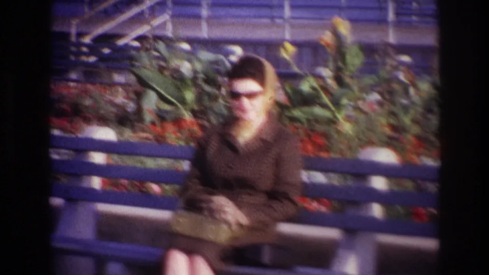 1967 Rockaway Beach frame of the woman facing the camera on a blue bench, with flowers, greenery, and railings behind her.