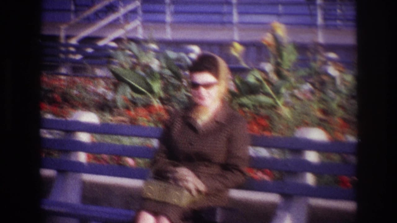 Adult woman in a yellow scarf, dark sunglasses, and brown coat sits on a blue bench in 1967 Rockaway Beach home-movie footage.