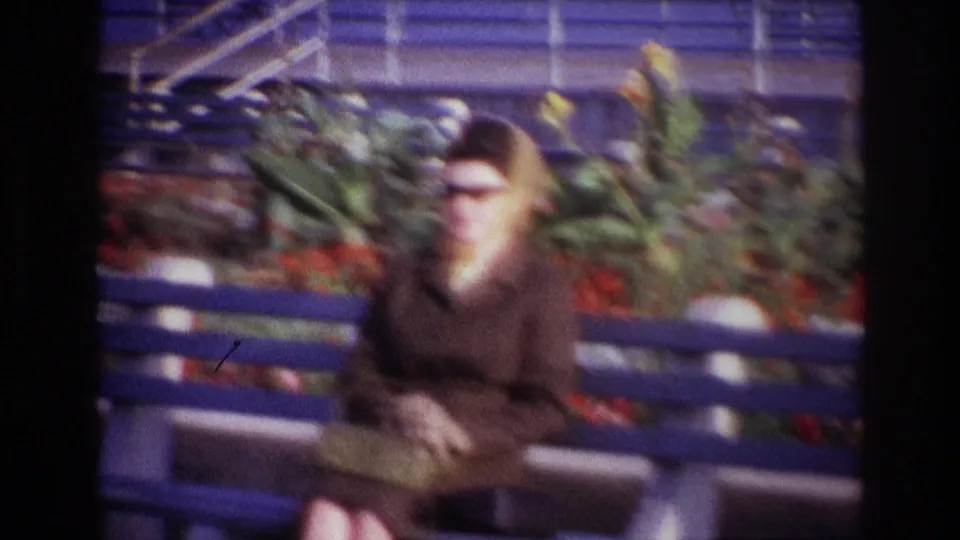The seated woman remains on the blue bench, slightly blurred by handheld 8mm motion, with garden planting behind her.