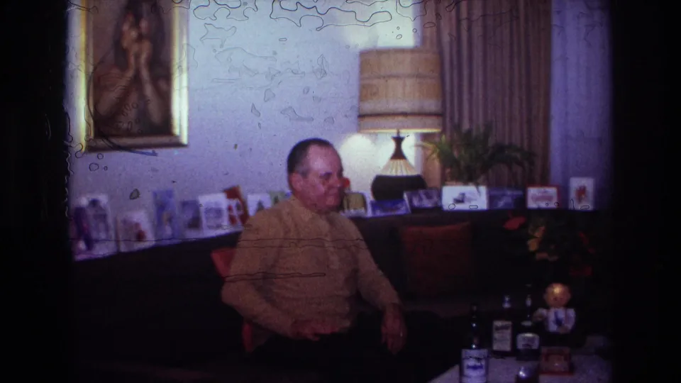 1967 home-movie frame from Rockaway Beach showing a man seated in a private living room with mid-century decor and tabletop objects.