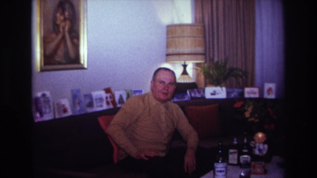 1967 Rockaway Beach home-movie frame showing a man in a brown sweater seated in a living room with cards, lamp, portrait, and bottles.