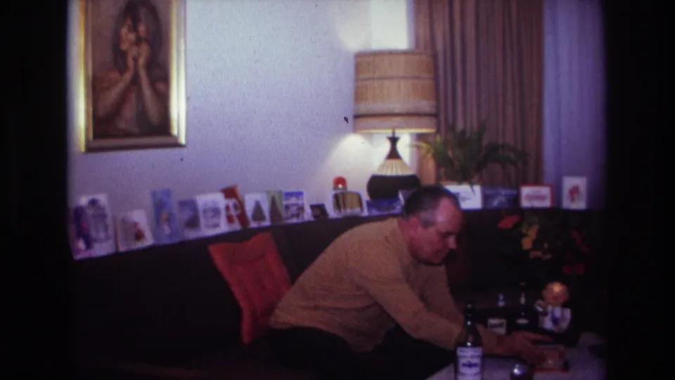 A home-movie frame shows the man bending toward a low table in a Rockaway Beach living room with holiday cards, lamp, plant, and framed portrait.