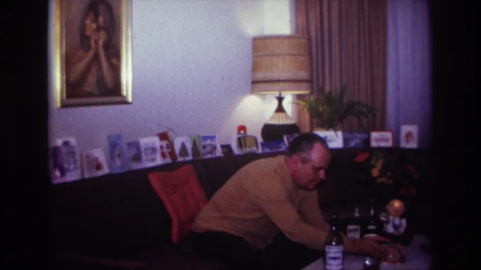 The final frame keeps the man leaning over the table in the 1967 Rockaway Beach living room, with cards, bottles, lamp, and portrait visible.